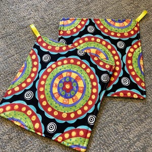 May include: Three colorful pot holders with a black background and a circular pattern. The pot holders have yellow fabric loops for hanging.