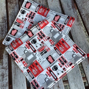 Puede incluir: Dos salvamanteles cuadrados y acolchados con temática de barbacoa. La tela presenta un patrón repetido de imágenes rojas, negras y blancas relacionadas con la barbacoa, incluyendo parrillas, utensilios y la palabra "BARBECUE".