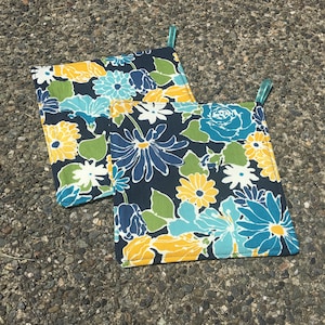 May include: Two square pot holders with a dark blue background and a floral pattern in shades of yellow, blue, green, and white. Each pot holder has a small loop for hanging.