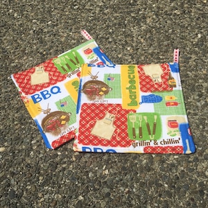May include: Two red, white, and blue kitchen pot holders with a BBQ theme. The pot holders have a variety of images including a grill, a spatula, a grill mitt, and the words "BBQ", "Let's eat!" and "grillin' & chillin'".