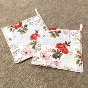 May include: Two square pot holders with a floral and butterfly pattern. They display red roses, pink butterflies, and green leaves on a white backdrop. Each pot holder has a pink loop for hanging.