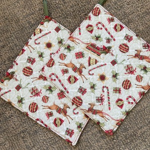 Puede incluir: Dos agarraderas de olla de algodón blanco con un tema navideño. Las agarraderas de olla presentan un fondo blanco con diseños navideños rojos y verdes que incluyen renos, bastones de caramelo, adornos y regalos. Las agarraderas de olla tienen lazos de tela verde para colgar.
