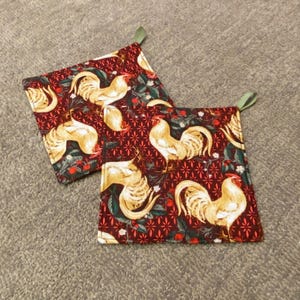 May include: Two square pot holders with a rooster pattern. The fabric is a deep red with a repeating pattern of roosters, flowers, and leaves. Each pot holder has a green loop for hanging.