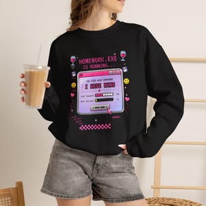 Op de afbeelding: Een zwart sweatshirt met lange mouwen en een grafische print van een computervenster met de tekst "HOMEWORK.EXE IS RUNNING..." en "THE KIDS HAVE HOMEWORK I HAVE WINE". Het ontwerp bevat voortgangsbalken, wijnglazen en een cursor.