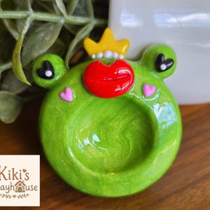 May include: A round, glossy, lime green frog-shaped trinket dish with a shimmering finish. The frog features a bright yellow crown, prominent red lips, black heart-shaped eyes, and small pink heart accents on its cheeks. The center is concave.