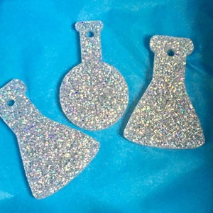 Puede incluir: Tres adornos plateados brillantes en forma de frasco con un agujero en la parte superior para colgar. Los adornos tienen diferentes formas: un círculo, un triángulo y una forma de frasco. Están sobre un fondo azul brillante.