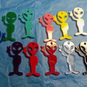 May include: A collection of colorful alien-shaped charms in various colors, including green, blue, red, pink, yellow, white, and silver. Each charm has a hole at the top and is waving with two fingers. The charms are displayed on a blue background.