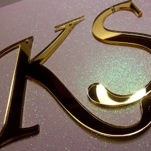 May include: Gold-colored mirrored acrylic letters 'K' and 'S' are displayed against a glittery, light-colored background. The letters are reflective, with a glossy finish, and appear to be freestanding. The 'K' has a classic serif design.
