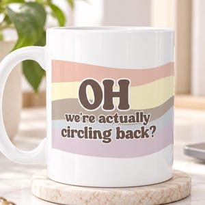 May include: A white ceramic mug featuring a wavy, multi-colored background in shades of peach, yellow, gray, and lavender. The text "OH we're actually circling back?" is printed in a bold, dark brown, rounded font across the center of the mug.