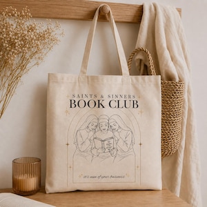 May include: A beige canvas tote bag featuring a black line art illustration of three nuns reading a book. The text reads SAINTS & SINNERS BOOK CLUB and it's nun of your business. The book title is 50 Shades of the Holy Spirit.