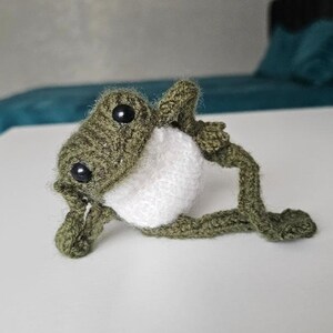 May include: A small, handmade crochet frog figurine crafted from olive green yarn with a contrasting white belly. The frog features two round, black plastic safety eyes and is posed in a reclining position with its limbs extended outward.