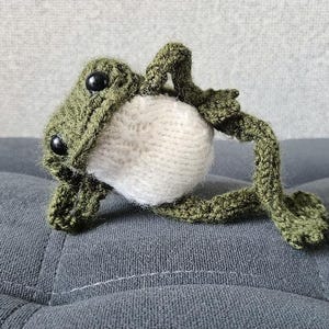 May include: A crocheted frog figurine crafted from dark olive green yarn with a contrasting cream-colored belly. The frog features two round, black plastic eyes and is posed in a reclining position with its limbs extended outward.