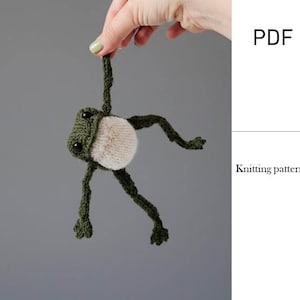 Könnte beinhalten: Ein dunkelgrüner gestrickter Frosch mit einem hellbeigen Bauch und schwarzen Perlenaugen. Der Frosch hat lange, dünne Gliedmaßen und hängt an einem Arm. Das Bild enthält den Text PDF und Knitting pattern, was auf eine digitale Strickanleitung hinweist.