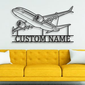 May include: A black metal wall art piece featuring a detailed outline of a commercial airplane in flight, positioned above a horizontal bar with the text CUSTOM NAME in bold, capital, sans-serif lettering, creating a clean, modern aesthetic.