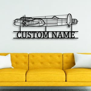 May include: A black metal wall art piece featuring a detailed outline of a multi-engine aircraft positioned above a horizontal bar with the text CUSTOM NAME in bold capital letters, mounted on a plain wall above a yellow sofa.