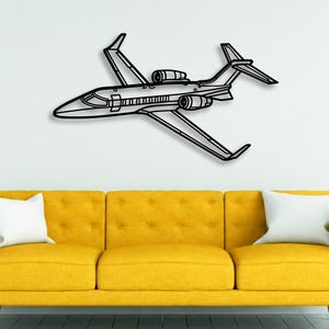 May include: A black metal wall art piece shaped like a private jet airplane. The design features a sleek fuselage, two wings, a T-tail stabilizer, and two jet engines mounted on the rear sides of the aircraft, rendered in a simple line art style.