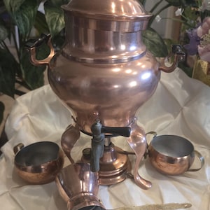 May include: A polished copper samovar featuring a central spigot with a black handle, two side handles, and a matching lid with a black knob. The set includes two small copper bowls, a copper cup, and a long, thin, decorative brass-toned metal spoon.