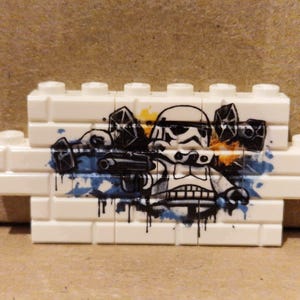 Könnte beinhalten: Ein weißer Kunststoff-Baustein-Verbund mit einem aufgedruckten Motiv eines Stormtrooper-Helms und TIE-Jägern. Das Design besteht aus schwarzen Linienzeichnungen mit blauen und gelben Farbspritzern, die einen Graffiti-Stil erzeugen.