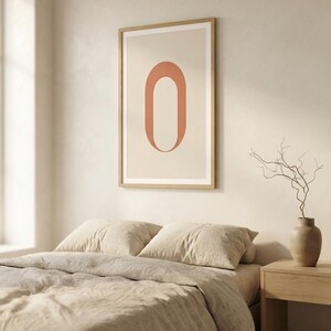 Op de afbeelding: Een rechthoekige wandkunstprint met een grote, verticaal geori&euml;nteerde, terracotta-kleurige letter O gecentreerd op een lichtbeige achtergrond. De print is ingelijst in een dunne, lichtgekleurde houten lijst en hangt aan een neutraal gekleurde muur.