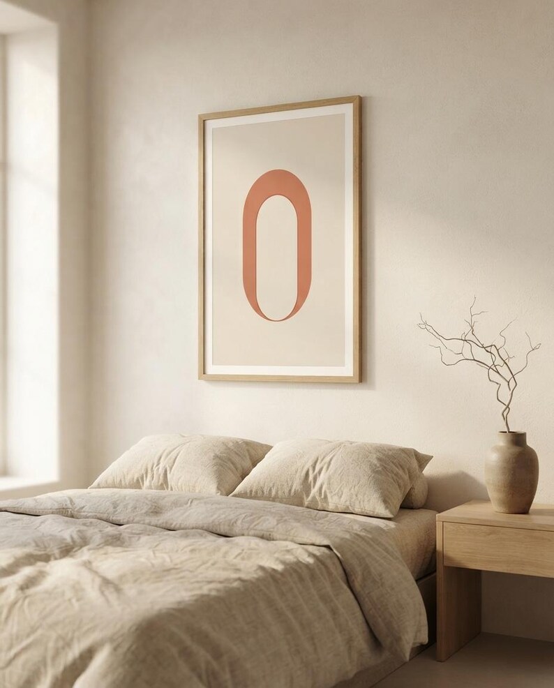 Op de afbeelding: Een rechthoekige wandkunstprint met een grote, verticaal geori&euml;nteerde, terracotta-kleurige letter O gecentreerd op een lichtbeige achtergrond. De print is ingelijst in een dunne, lichtgekleurde houten lijst en hangt aan een neutraal gekleurde muur.