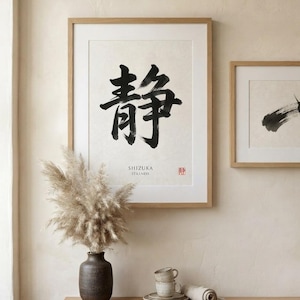 A framed wall art print featuring large black Japanese calligraphy characters, the text SHIZUKA STILLNESS, and a small red seal. The print is housed in a light wood frame, displayed alongside a second smaller framed abstract brush stroke.
