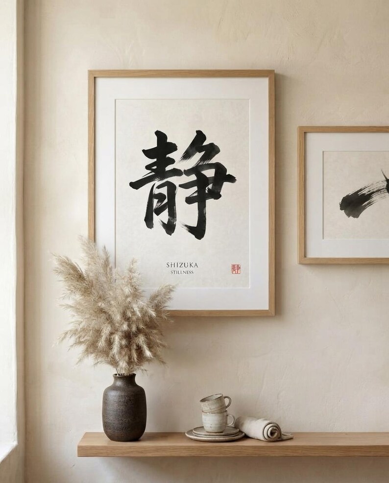 A framed wall art print featuring large black Japanese calligraphy characters, the text SHIZUKA STILLNESS, and a small red seal. The print is housed in a light wood frame, displayed alongside a second smaller framed abstract brush stroke.