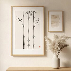 A large framed wall art print featuring a black ink illustration of three bamboo stalks on a cream background, accompanied by a smaller framed abstract watercolor print and a white ceramic vase with dried pampas grass.