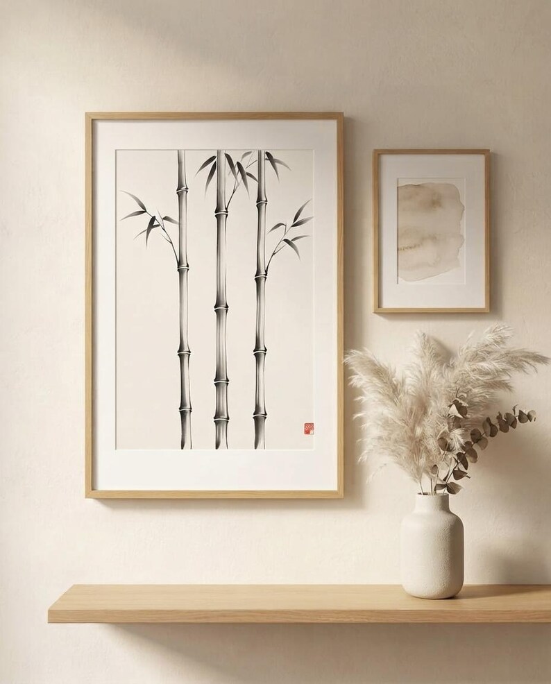 A large framed wall art print featuring a black ink illustration of three bamboo stalks on a cream background, accompanied by a smaller framed abstract watercolor print and a white ceramic vase with dried pampas grass.