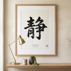 A framed wall art print featuring large black Japanese calligraphy characters on a textured off-white background. Below the characters, the text reads SHIZUKA STILLNESS, accompanied by a small red square stamp in the bottom right corner.