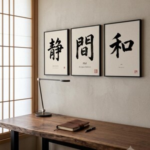 Op de afbeelding: Drie ingelijste wandkunstwerken met zwarte Japanse kalligrafiekarakters op een witte achtergrond. Elke print bevat kleine Engelse tekst en een rode stempel. De getoonde karakters zijn 静, 間 en 和, wat staat voor stilte, de ruimte tussen en harmonie.