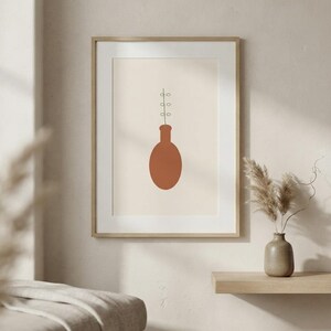 May include: A framed wall art print featuring a minimalist illustration of a terracotta-colored oval vase with a thin black stem and small circular leaves extending upward from the opening, set against a solid light beige background.