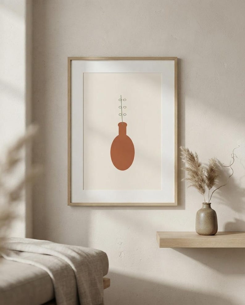 May include: A framed wall art print featuring a minimalist illustration of a terracotta-colored oval vase with a thin black stem and small circular leaves extending upward from the opening, set against a solid light beige background.