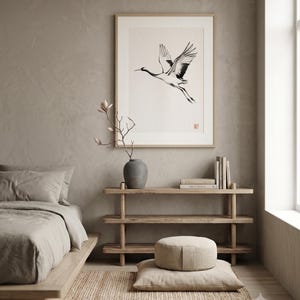 Sumi-e crane print framed in oak, seen on three quiet walls — above a linen bed, on a gallery wall and above an oak console.