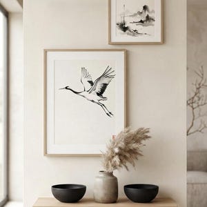 Two framed wall art prints featuring black and white ink-style illustrations of a flying crane and a landscape scene, displayed above a wooden shelf holding a ceramic vase with dried pampas grass and two matte black bowls.