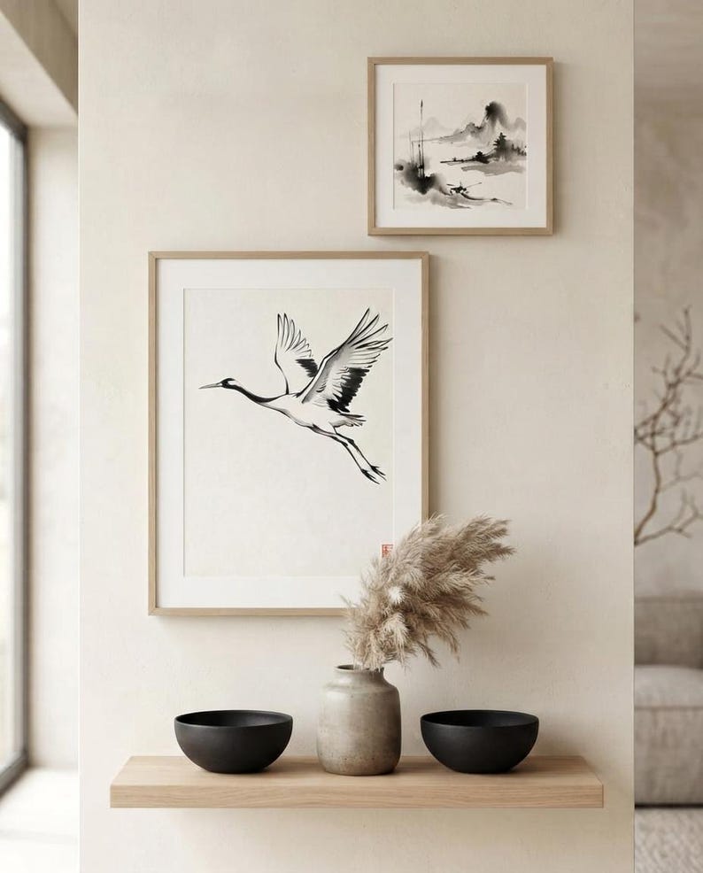 Two framed wall art prints featuring black and white ink-style illustrations of a flying crane and a landscape scene, displayed above a wooden shelf holding a ceramic vase with dried pampas grass and two matte black bowls.