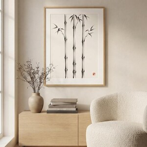 A framed wall art print featuring a black and white ink wash illustration of three bamboo stalks with leaves, set against a cream background, with a small red square seal mark in the lower right corner of the artwork.