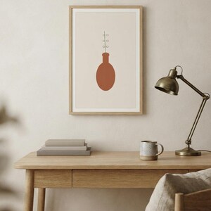 May include: A framed wall art print featuring a solid terracotta-colored oval vase shape centered on a light beige background. A thin black line extends upward from the vase, displaying three pairs of small, simple, outlined circular leaves.