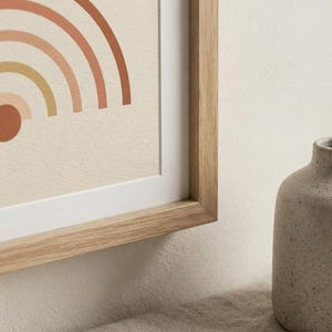 May include: A framed print featuring a partial graphic of a rainbow with concentric arches in shades of terracotta, peach, and mustard yellow on a cream background, encased in a light-toned wooden frame with a white mat border.