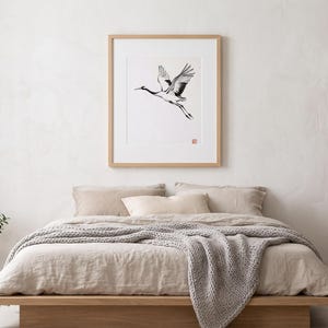Sumi-e crane print framed in oak, seen on three quiet walls — above a linen bed, on a gallery wall and above an oak console.