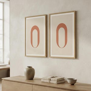 Op de afbeelding: Twee ingelijste verticale wandkunstprints met een gecentreerde terracotta-kleurige ovale vorm op een lichtbeige achtergrond. De lijsten zijn gemaakt van lichtgekleurd hout en de prints hangen naast elkaar aan een muur boven een houten console.