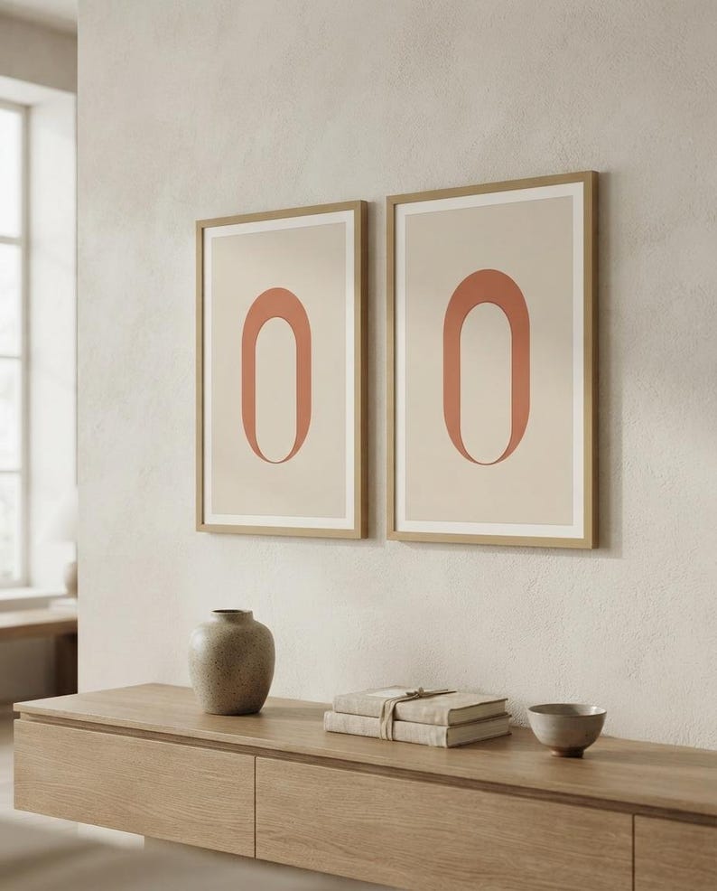 Op de afbeelding: Twee ingelijste verticale wandkunstprints met een gecentreerde terracotta-kleurige ovale vorm op een lichtbeige achtergrond. De lijsten zijn gemaakt van lichtgekleurd hout en de prints hangen naast elkaar aan een muur boven een houten console.