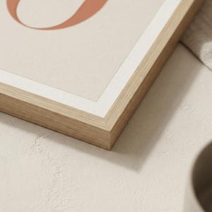 Op de afbeelding: Een ingelijste wandkunstprint met een deel van een grote, terracotta-kleurige letter U tegen een lichtbeige achtergrond. Het kunstwerk zit in een lichtgekleurde houten lijst met een dunne witte rand rondom de print.