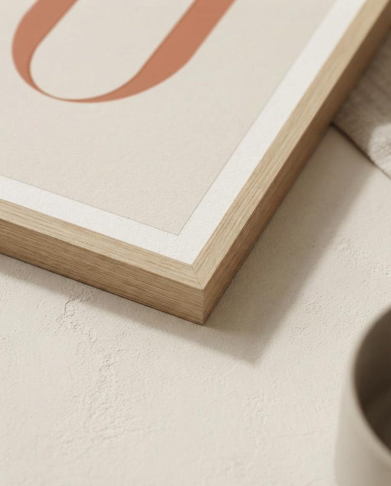 Op de afbeelding: Een ingelijste wandkunstprint met een deel van een grote, terracotta-kleurige letter U tegen een lichtbeige achtergrond. Het kunstwerk zit in een lichtgekleurde houten lijst met een dunne witte rand rondom de print.