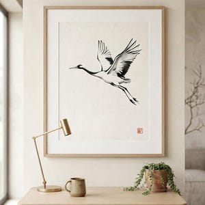 A framed wall art print featuring a black and white ink wash style illustration of a crane in flight. The bird is depicted with extended wings and long legs against a cream background, with a small red square seal mark in the corner.