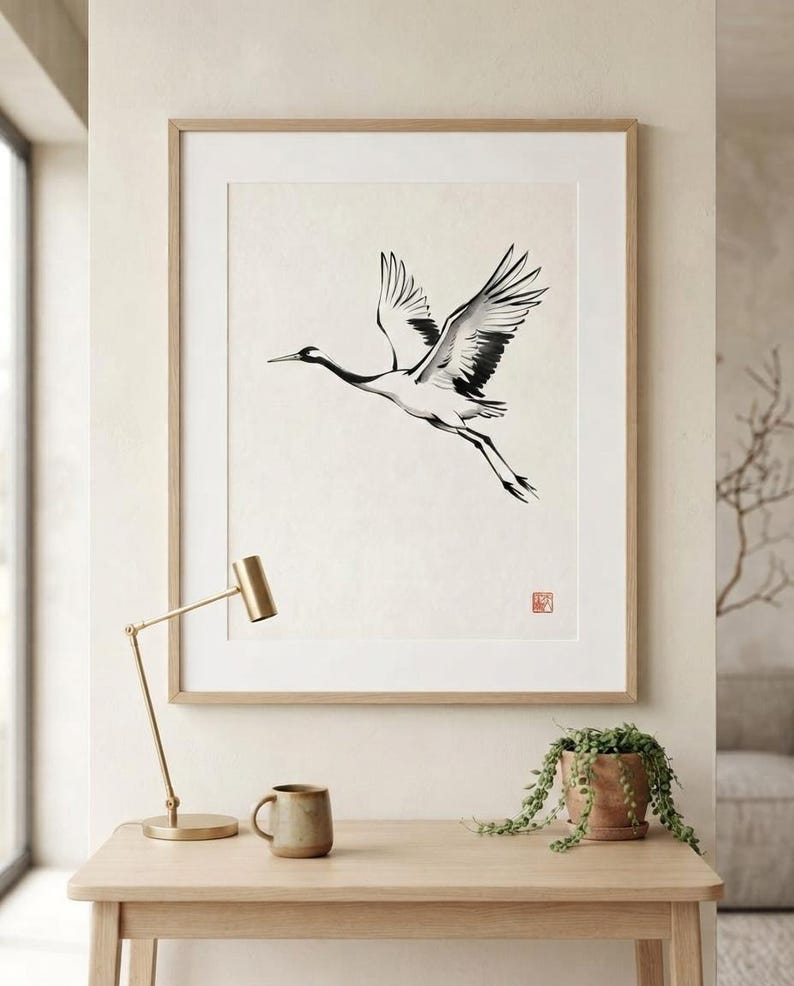 A framed wall art print featuring a black and white ink wash style illustration of a crane in flight. The bird is depicted with extended wings and long legs against a cream background, with a small red square seal mark in the corner.