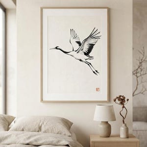 Sumi-e crane print framed in oak, seen on three quiet walls — above a linen bed, on a gallery wall and above an oak console.