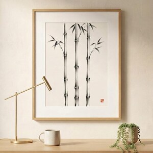 A framed wall art print featuring a black ink illustration of three bamboo stalks on a cream background. The artwork includes a small red square seal in the lower right corner. The print is encased in a light-colored wooden frame.