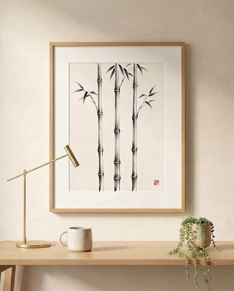A framed wall art print featuring a black ink illustration of three bamboo stalks on a cream background. The artwork includes a small red square seal in the lower right corner. The print is encased in a light-colored wooden frame.