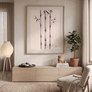 Pode incluir: Uma impress&atilde;o de arte emoldurada com uma ilustra&ccedil;&atilde;o a tinta preta de tr&ecirc;s caules de bambu num fundo bege claro. A obra de arte &eacute; apresentada numa moldura de madeira clara e est&aacute; pendurada centrada numa parede sobre um m&oacute;vel de madeira.