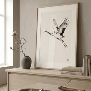 Sumi-e crane print framed in oak, seen on three quiet walls — above a linen bed, on a gallery wall and above an oak console.